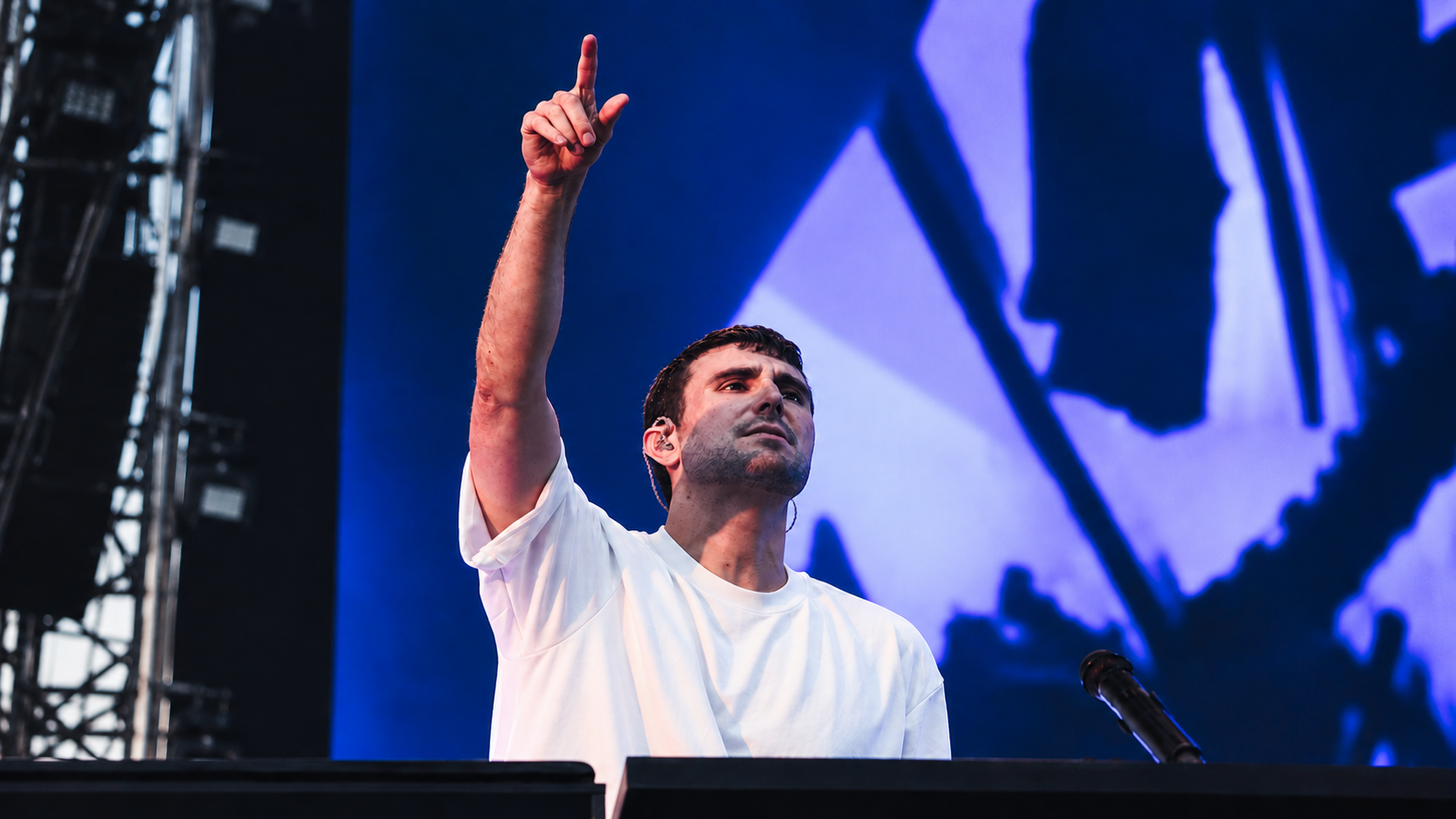 Fred again.. performing on stage with one arm raised in front of a blue screen