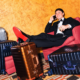ohn Summit sitting in a red chair surrounded by aluminum suitcases for his Away partnership campaign