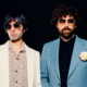 ustice duo posing in sunglasses against a black background, wearing white and blue suits