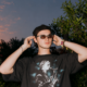 Mau P wearing sunglasses and a black cap outdoors at sunset