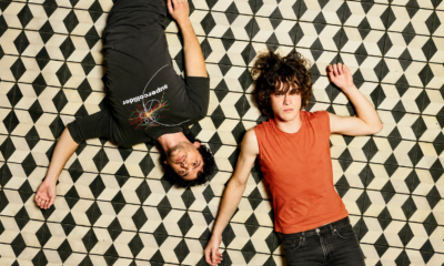 MGMT lying on a black-and-white geometric tiled floor, one wearing black and the other an orange sleeveless top