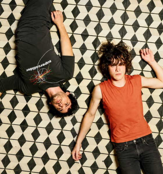 MGMT lying on a black-and-white geometric tiled floor, one wearing black and the other an orange sleeveless top