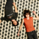 MGMT lying on a black-and-white geometric tiled floor, one wearing black and the other an orange sleeveless top