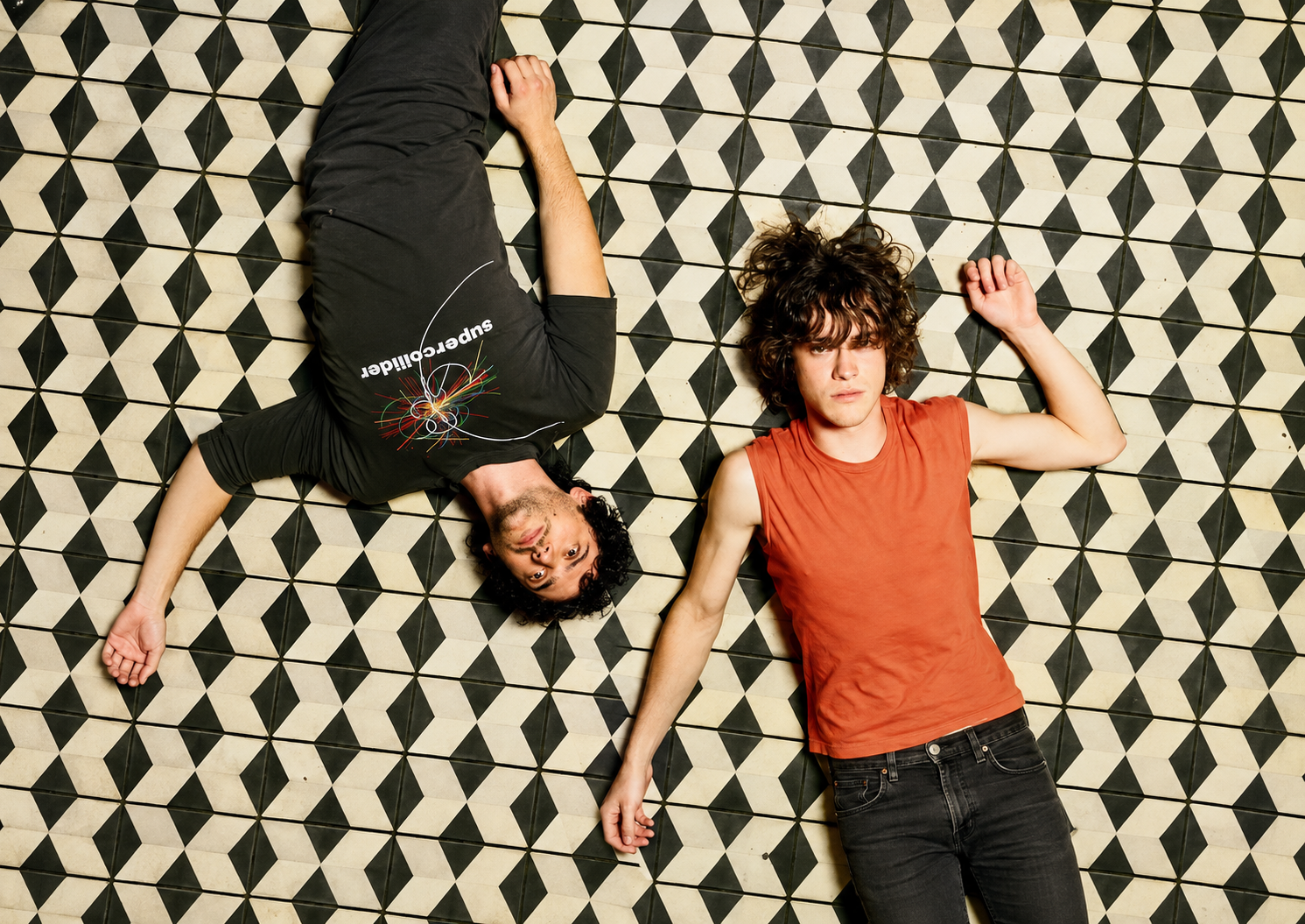 MGMT lying on a black-and-white geometric tiled floor, one wearing black and the other an orange sleeveless top