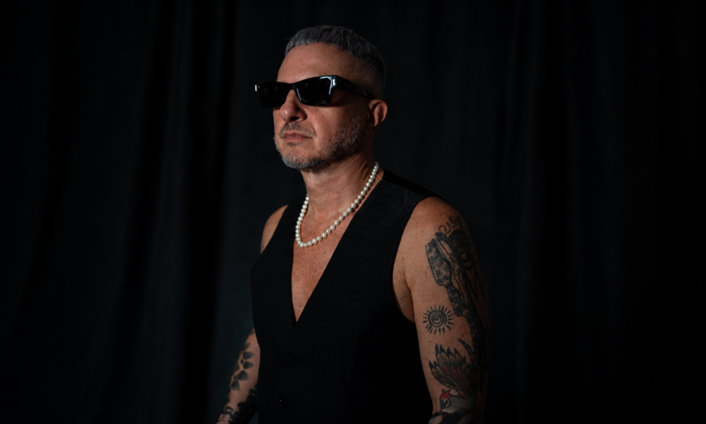 MoBlack wearing black sunglasses and a sleeveless black top against a dark background.