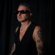 MoBlack wearing black sunglasses and a sleeveless black top against a dark background.