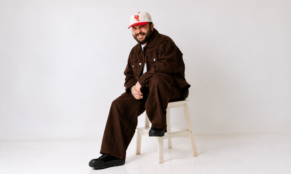 Moojo sitting on a white chair wearing a brown outfit and white cap against a light studio background