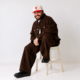 Moojo sitting on a white chair wearing a brown outfit and white cap against a light studio background