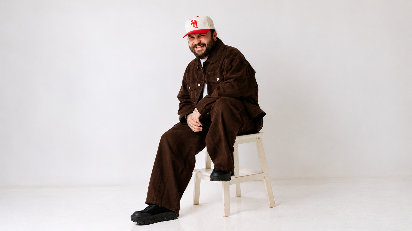 Moojo sitting on a white chair wearing a brown outfit and white cap against a light studio background