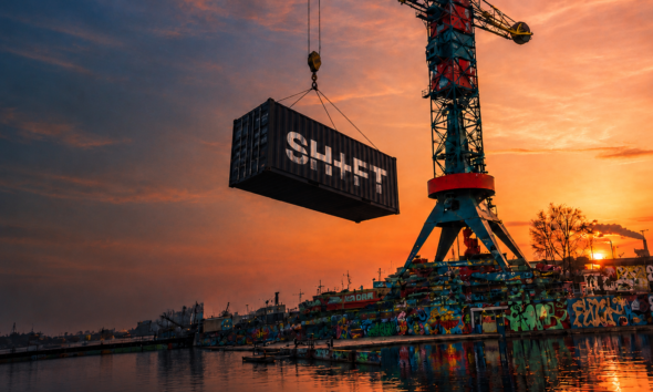 A SHIFT-branded shipping container suspended from a crane above a graffiti-covered waterfront at sunset.