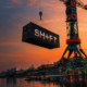 A SHIFT-branded shipping container suspended from a crane above a graffiti-covered waterfront at sunset.