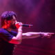 Skrillex performing on stage with a microphone under red and purple lighting.