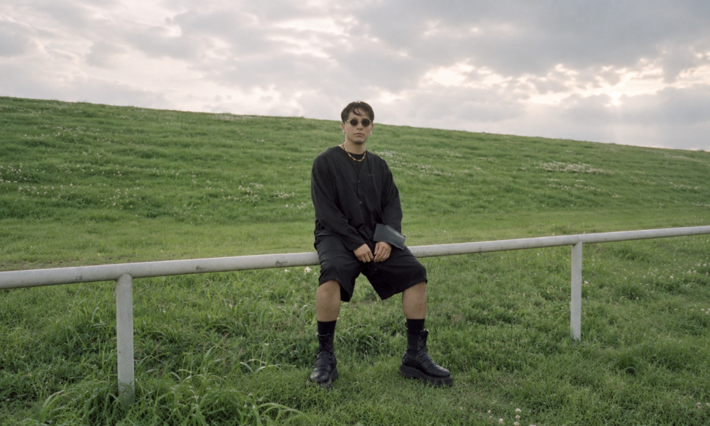 WhereIsFenix seated outdoors in a grassy field wearing black clothing and sunglasses for his debut album Curiosity