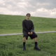 WhereIsFenix seated outdoors in a grassy field wearing black clothing and sunglasses for his debut album Curiosity