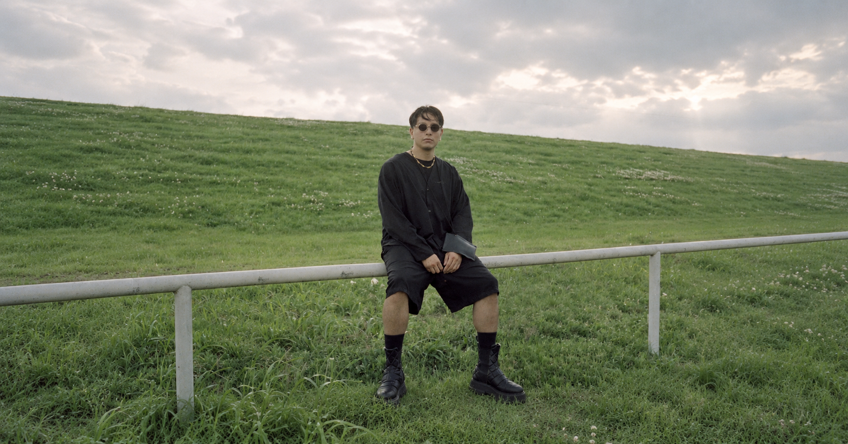WhereIsFenix seated outdoors in a grassy field wearing black clothing and sunglasses for his debut album Curiosity