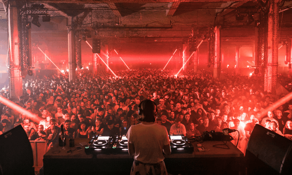 View from behind the decks as a DJ plays to a packed crowd under red lasers at The Warehouse Project