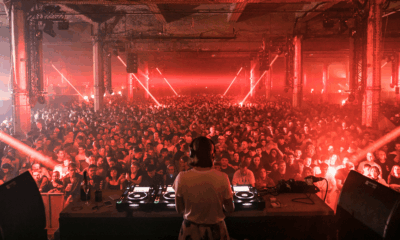 View from behind the decks as a DJ plays to a packed crowd under red lasers at The Warehouse Project
