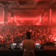 View from behind the decks as a DJ plays to a packed crowd under red lasers at The Warehouse Project